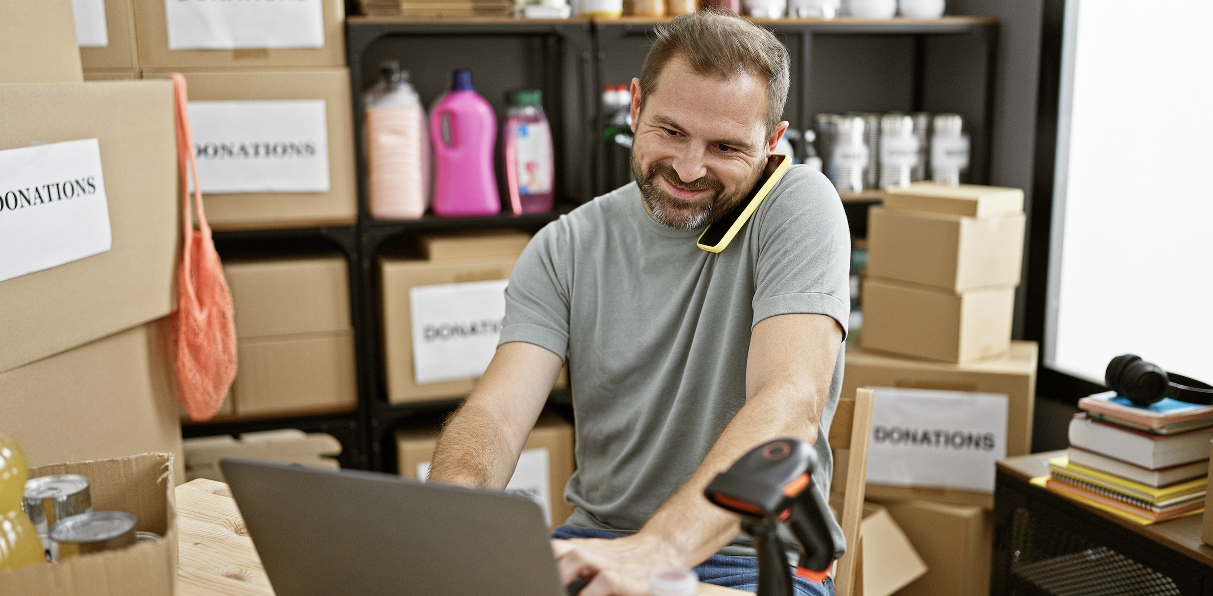 Middle-aged man multitasking on a laptop with a phone at a busy donation center warehouse.